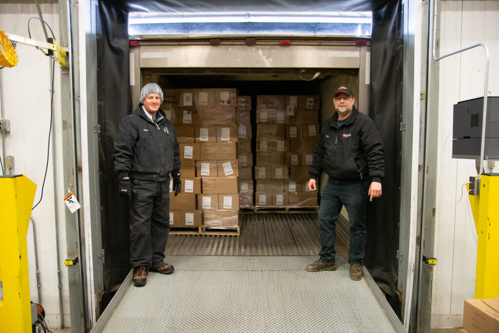 Two men stand in the back of an open refrigerated trailer of cheeses